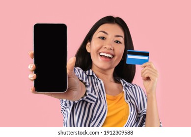 Online Shopping. Portrait Of Cheerful Young Asian Woman Holding Credit Card In Hand And Showing Mobile Phone With Black Blank Screen To Camera. Gadget With Empty Free Space For Mock Up, Digital Pay