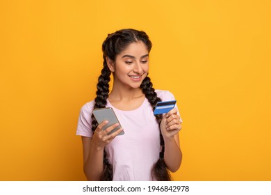 Online Shopping. Portrait Of Smiling Indian Woman Using Her Mobile Phone And Holding Debit Credit Card Isolated Over Yellow Studio Background. Money Transfer, Digital Instant Payment Concept