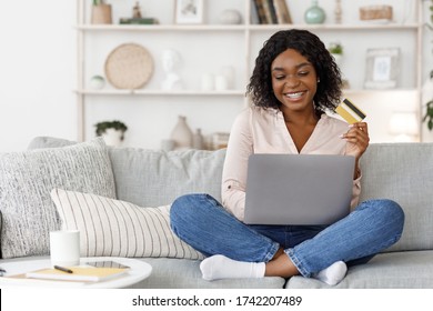 Online Shopping. Positive Young Black Girl Using Laptop and Credit Card At Home, Making Purchases In Internet, Sitting On Couch In Living Room
