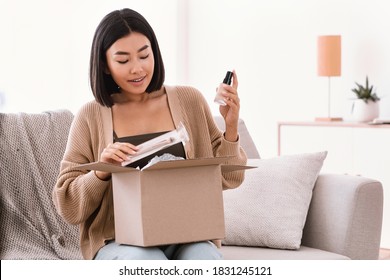 Online Shopping. Smiling Asian Woman Opening Parcel Box With Cosmetics At Home, Holding Makeup Products, Unpacking Gift Sitting On The Couch At Home. Lady Satisfied With Delivery Service