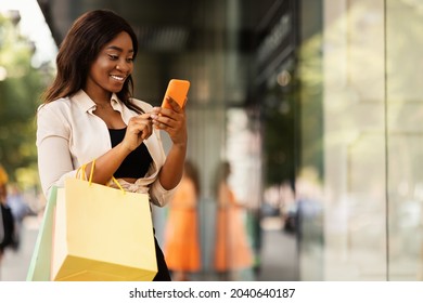 Online Shopping. Smiling Black Woman Using Her Mobile Phone, Holding Shopper Bags, Standing Outdoors Near Mall Window, Choosing Clothes And Presents, Taking Photo, Free Space. Special Offer