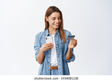 Online Shopping. Smiling Good-looking Woman Paying For Order, Using Plastic Credit Card To Pay With Mobile Phone, Standing Against White Background