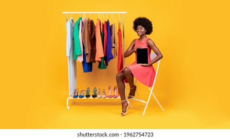 Online Shopping. Stylish African American Woman Showing Digital Tablet Blank Screen Recommending Clothing Store Website Sitting Near Rack With Colorful Female Clothes Over Yellow Background. Panorama - Powered by Shutterstock