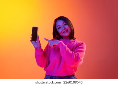 Online shopping  Young girl in pink sweater smiling  pointing phone screen over gradient orange background in neon light  Concept emotions  facial expression  youth  inspiration  sales  ad