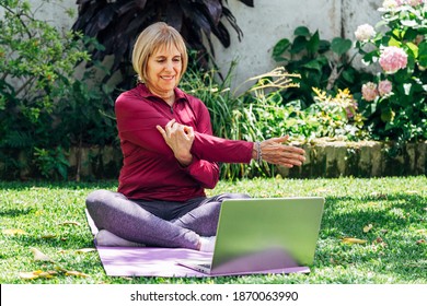 Online sport fitness yoga training workout. Senior woman in the garden of her home doing exercises over a rubber mat and watching the laptop. Coach in video conference. Healthy lifestyle concept.