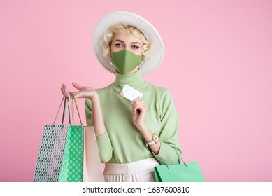 Online Spring Shopping During Quarantine Conception: Fashionable Woman Wearing Protective Mask Posing With Colorful Paper Bags And Plastic Bank Card. Pink Background. Copy, Empty Space For Text