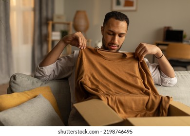 Online Store And Delivery, Remote Shopping Concept. Handsome Young Black Guy Unboxing Cardboard Parcel, Trying On Delivered Purchase, Taking Out New Clothes At Home