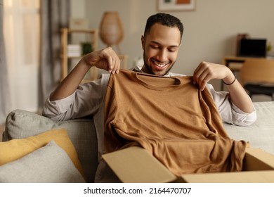 Online Store And Delivery, Successful Shopping Concept. Cheerful Black Guy Unboxing Cardboard Parcel, Satisfied With Great Purchase, Taking Out New Clothes At Home