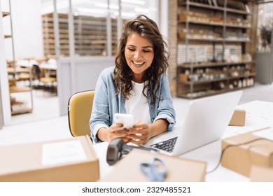 Online store owner reading a text message on her cellphone. Happy businesswoman making plans for product shipping in her warehouse. Female entrepreneur running an e-commerce small business.