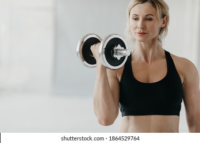 Online Strength Training At Home With Trainer During Lockdown And Self-isolation. Portrait Of Calm Muscular Adult Female Or Coach In Bra Doing Exercises With Dumbbells In Loft Interior Of Home Or Gym