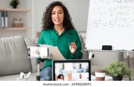 Online Studying, Distance Learning, Virtual Class. Young female teacher shooting lesson on smart phone camera, sitting at desk near flipchart, talking to pc screen with group of diverse students - Powered by Shutterstock