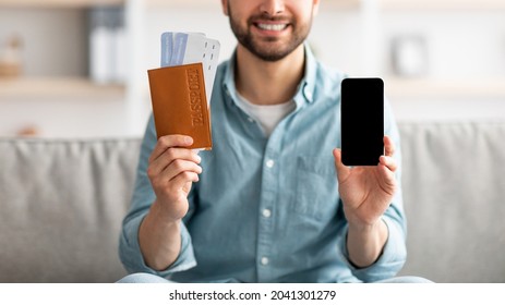 Online tour ot hotel booking app. Young guy holding passport and tickets, demonstrating smartphone with mockup screen at home. Millennial man advertising internet tourist agency