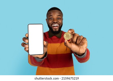 Online Trading. Excited Black Man Showing Golden Bitcoin Coin And Blank Smartphone With White Screen At Camera, African American Guy Advertising Crypto App While Posing On Blue Background, Mockup