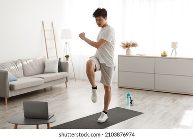 Online Training, Home Workout. Focused Sporty Asian Guy Standing In Living Room Doing Twist Knees To Elbow Exercise Using Laptop Pc. Domestic Male Fitness And Sporty Lifestyle