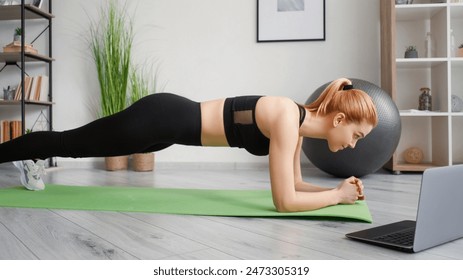 Online training. Morning workout. Balance exercise. Healthy lifestyle. Fit woman activewear standing in plank on yoga mat with laptop at home interior side view. - Powered by Shutterstock