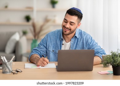 Online Training. Portrait Of Happy Jewish Man In Kipa Working Or Studying Remotely At Home Office, Writing In His Notebook With Pen, Taking Notes, Using Laptop And Sitting At Table. E-learning