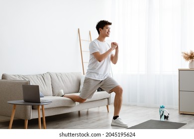 Online Training. Smiling Sporty Asian Guy Doing Single-Leg Split Squats Near Couch Exercising Using Laptop Computer During Workout At Home. Domestic Male Fitness And Sporty Lifestyle. Side View