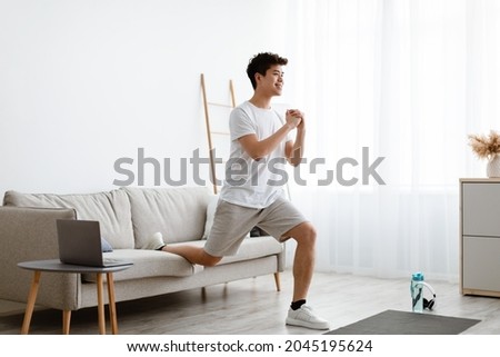 Online Training. Smiling Sporty Asian Guy Doing Single-Leg Split Squats Near Couch Exercising Using Laptop Computer During Workout At Home. Domestic Male Fitness And Sporty Lifestyle. Side View