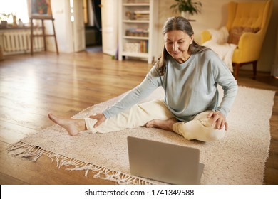 Online Training, Technology, Health And Activity. Happy Active Barefoot Mature Woman Sitting On Floor In Front Of Open Laptop, Doing Yoga, Repeating Exercises After Professional Instructor On Video