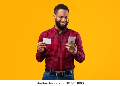 Online Wallet Concept. Smiling Black Man Showing Credit Card, Holding Smart Phone Isolated Over Yellow Studio Background Wall. Satisfied African American Guy Shopping, Buying Via Internet