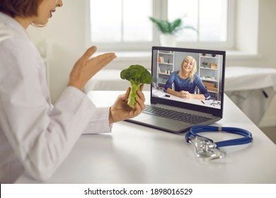 Online Wellness Consultation Via Video Call: Doctor, Dietitian Or Nutritionist Sitting At Laptop, Holding Broccoli, Talking To Senior Patient And Giving Her Tips On Happy Long Life And Healthy Diet