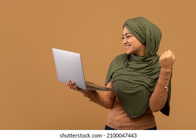 Online win. Excited muslim lady in hijab holding laptop and celebrating success with raised fist, standing over beige background, free space
