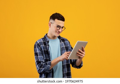 Online work, communiction, education. Cool young man browsing web on tablet pc over orange studio background. Funky student in glasses using touch pad, watching videos, posting in social media