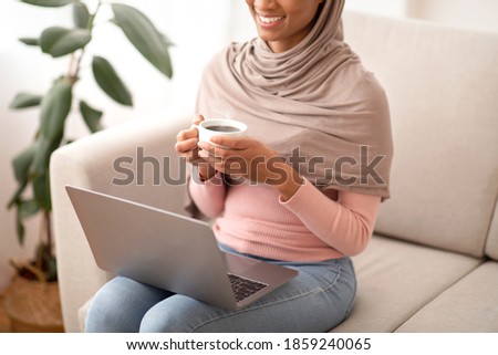 Online work, education, communication. Unrecognizable black woman in hijab using laptop and drinking coffee at home. Young Muslim lady having web lesson or conference indoors
