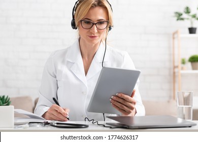 Online Work With Patients. Busy Woman Doctor In Headphones Holds Tablet In Hands And Makes Notes In Notebook In Interior Of Office