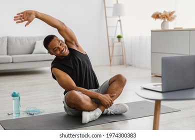 Online Workout. Cheerful Athletic African American Guy Doing Stretching Exercise At Laptop Training Sitting On Gymnastics Mat In Living Room At Home. Male Fitness Routine Concept