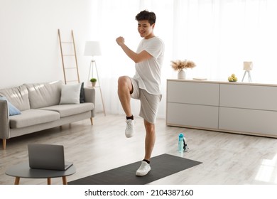 Online Workout Concept. Cheerful Smiling Asian Guy Training In Living Room, Doing Twist Knees To Elbow Exercise. Happy Man Standing And Lifting Leg Up, Using Laptop Watching Tutorial Lookign At Screen