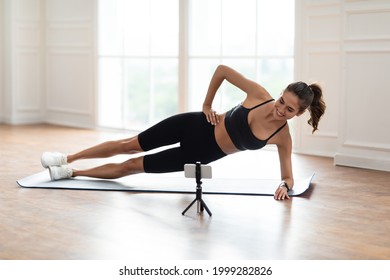 Online Workout. Smiling Sporty Female Standing In Side Plank Position On Yoga Mat Near Window, Looking At Cellphone On Tripod, Watching Tutorial Or Recording Video For Blog, Having Chat With Coach