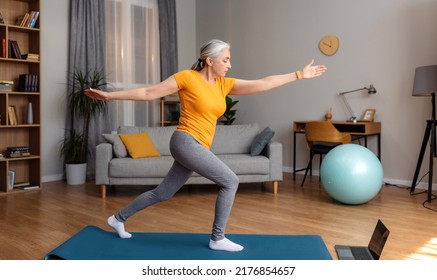 Online Yoga Training. Active Senior Woman Exercising In Front Of Laptop, Standing On Fitness Mat, Watching Video On Internet Or Having Online Yoga Class At Home