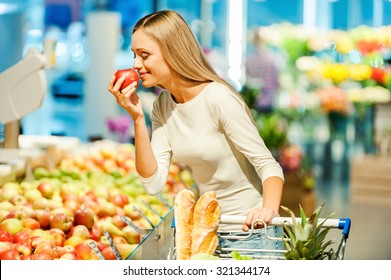 Only The Best Fruits And Vegetables. Beautiful Young Woman Holding Apple And Smelling It With Smile While Standing In A Food Store 