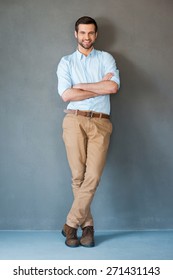 Only Positive Emotions. Full Length Of Handsome Young Man In Shirt Keeping Arms Crossed And Smiling At Camera While Standing Against Grey Background 