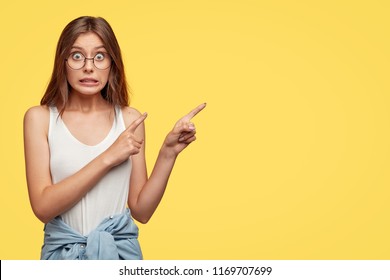 Oops, come and look there! Lovely young woman feels worried and ashamed, points with both index fingers at upper right corner, isolated over yellow background, makes mistake and looks awkward