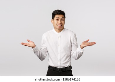 Oops sorry but cant help. Indecisive cute asian young man, businessman dont know how situation happened, explain himself, shrugging and smiling awkward, standing unsure white background