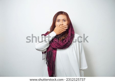 Oops! Surprised young Asian Muslim woman covering mouth with hands and staring at camera while standing against white background