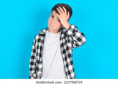 Oops, What Did I Do? Caucasian Kid Boy Wearing Plaid Shirt Over Blue Background Holding Hand On Forehead With Frightened And Regret Expression.