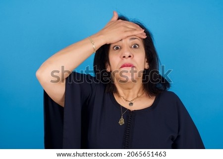 Oops, what did I do? middle aged Arab woman standing against blue background holding hand on forehead with frightened and regret expression.