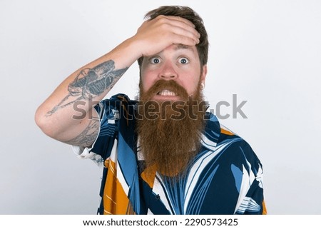 Oops, what did I do? red haired man wearing printed shirt over white studio background holding hand on forehead with frightened and regret expression.