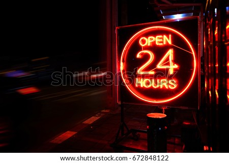 Open 24 Hours neon sign on the road side at night with moving cars.