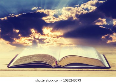 Open Bible On Wooden Table Over The Light Through Dark Cloudy Sunset Sky. Christian Concept Show The Word Of God Is The Light Of Mandkind