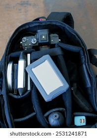An open black camera backpack filled with photography equipment, including a camera, lens, hard drive, and other accessories. The camera is visible in the top compartment.