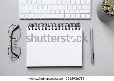 Open blank notebook, computer keyboard, glasses, pen and floral decor on light grey background, flat lay