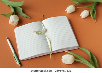 Open blank notebook with lined pages surrounded by white tulips, green leaves, and a pen on an orange background.