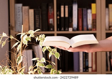 an open book held in one hand, next to a table with a bookcase in the background.