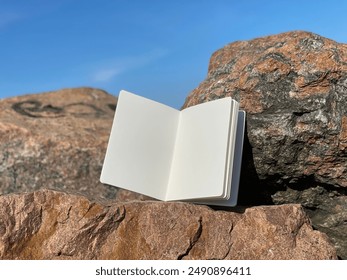 Open book, notebook or diary lies on the stones, sunny summer day. Mockup photo with empty white pages for text or advertising. Blue sky, rough rocks and stones.