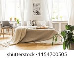 Open book on a gray, wooden armchair by a cozy bed with breakfast tray in a stylish bedroom interior with natural light coming through big windows
