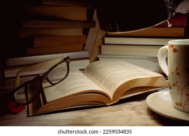 open book with a pair of glasses on top of it in the light of a lamp, a cup and in the background books piled up - Powered by Shutterstock